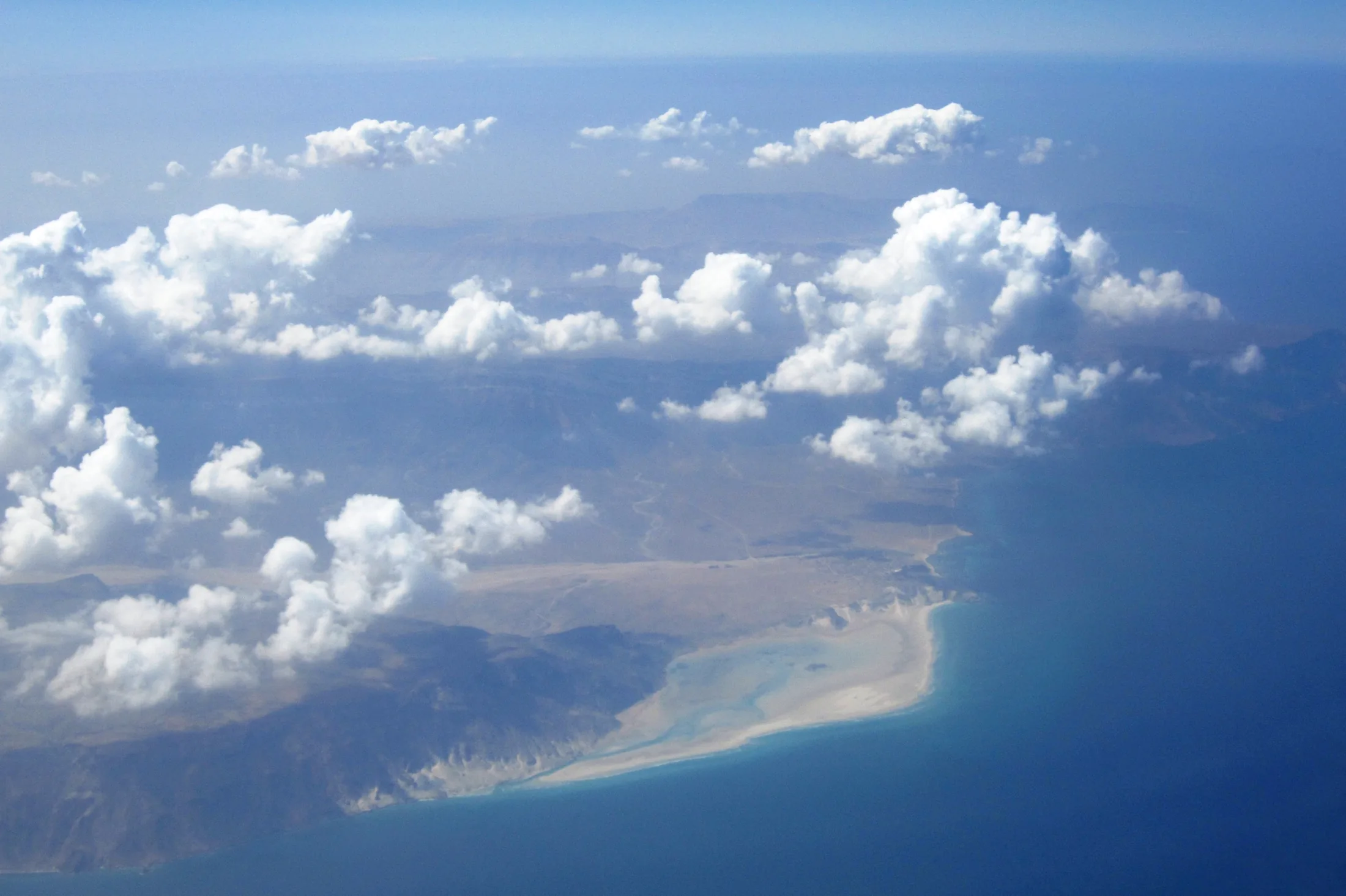 Aerial view of Detwah Lagoon’s pale sandy shoreline and shallow turquoise water, with clouds over Socotra’s coast.